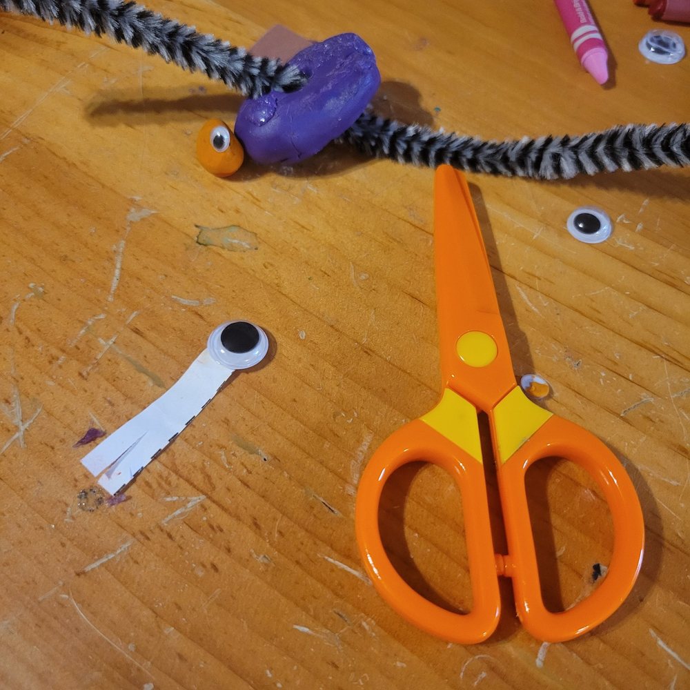 The original Tricky Eye: a slip of white scrap paper with a googly eye, on a wooden craft table beside kid scissors, clay and pipe cleaners